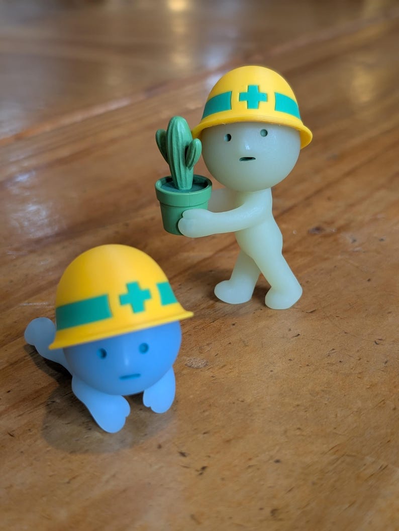 Puede incluir: Dos peque&ntilde;as figuras, una azul y otra verde p&aacute;lido, ambas con cascos amarillos con rayas verdes y una cruz blanca. La figura verde sostiene un peque&ntilde;o cactus verde en maceta. La figura azul est&aacute; en el suelo.