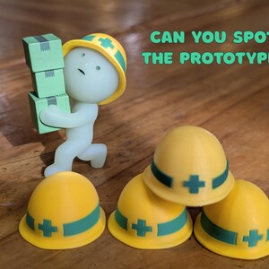 Puede incluir: Una figura caprichosa de un trabajador que lleva cajas verdes, con un casco amarillo con una banda y una cruz verdes. La imagen tambi&eacute;n muestra tres cascos amarillos con bandas y cruces verdes. Texto: "CAN YOU SPOT THE PROTOTYPE?"