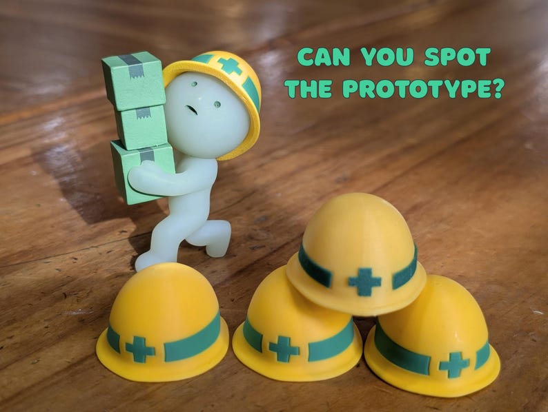 Puede incluir: Una figura caprichosa de un trabajador que lleva cajas verdes, con un casco amarillo con una banda y una cruz verdes. La imagen tambi&eacute;n muestra tres cascos amarillos con bandas y cruces verdes. Texto: "CAN YOU SPOT THE PROTOTYPE?"