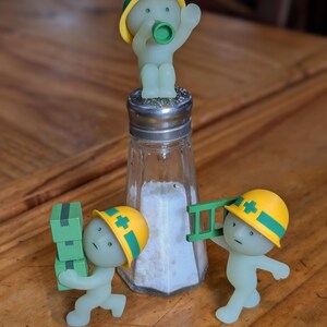 Puede incluir: Un salero novedoso con un cuerpo de vidrio transparente y una tapa de metal, lleno de sal. Tres peque&ntilde;as figuras verdes brillantes que llevan cascos de construcci&oacute;n amarillos est&aacute;n colocadas alrededor del salero, una encima, otra llevando cajas y otra con una escalera.
