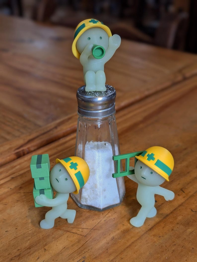 Puede incluir: Un salero novedoso con un cuerpo de vidrio transparente y una tapa de metal, lleno de sal. Tres peque&ntilde;as figuras verdes brillantes que llevan cascos de construcci&oacute;n amarillos est&aacute;n colocadas alrededor del salero, una encima, otra llevando cajas y otra con una escalera.
