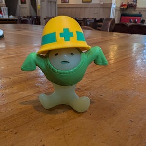 Puede incluir: Una figura caprichosa que representa a un trabajador de la construcci&oacute;n. Tiene un casco amarillo con una cruz verde, brazos verdes y un cuerpo de color claro. La figura est&aacute; sobre una mesa de madera.