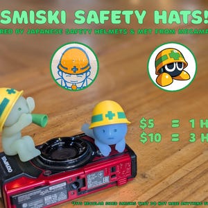 Puede incluir: &iexcl;Sombreros de seguridad Smiski! Dos peque&ntilde;as figuras que llevan cascos de seguridad amarillos con detalles verdes, inspirados en los cascos de seguridad japoneses. El texto dice "Smiski Safety Hats!" y "Inspired by Japanese Safety Helmets & Met from Megaman."