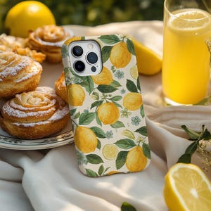 May include: A phone case with a lemon pattern rests on a white cloth, next to a glass of juice and pastries. The case features yellow lemons, green leaves, and small blue flowers. A sliced lemon and whole lemons are also visible.