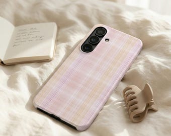 May include: A pink and white plaid phone case with a triple-lens camera is displayed on a bed. A small, open notebook and a beige hair clip are also present. The scene is set on a soft, textured surface.