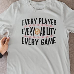 May include: A light gray t-shirt with the text "EVERY PLAYER, EVERY ABILITY, EVERY GAME" in black. The words are separated by pink baseball stitching and a baseball glove graphic.