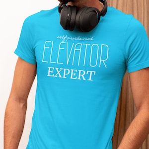 May include: A turquoise blue t-shirt with the words "self proclaimed ELEVATOR EXPERT" printed in white. The shirt is short-sleeved and has a crew neck. Black headphones are around the neck.