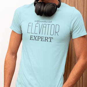 May include: Light blue t-shirt with the text "self proclaimed ELEVATOR EXPERT" in dark gray. The shirt is short-sleeved and has a crew neck. Black headphones are around the neck.