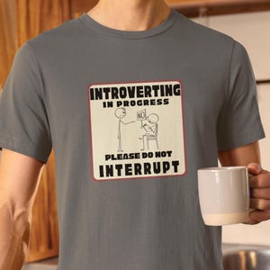 May include: A gray t-shirt with a white sign that says "Introverting in Progress. Please do not interrupt." The sign has a stick figure holding a sign that says "No" and another stick figure sitting down with a pen and paper.