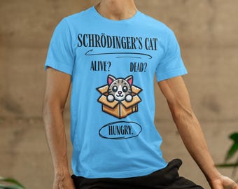 Op de afbeelding: Een lichtblauw t-shirt met een cartoonkat in een doos. De tekst op het shirt luidt "Schrödinger's Cat" met een lijn eronder en de woorden "Alive? Dead?" onder de lijn. De tekst "Hungry." staat in een cirkel onder de kat.