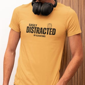 May include: Mustard yellow t-shirt with the text "EASILY DISTRACTED BY ELEVATORS" in black. The shirt features an elevator graphic. Black headphones are around the neck.