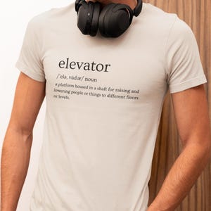 May include: A light grey t-shirt with a definition of the word "elevator" printed on the front. The definition reads:  /'elə, vädər/noun a platform housed in a shaft for raising and lowering people or things to different floors or levels.