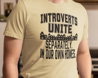 May include: A beige t-shirt with black text that reads "Introverts Unite Separately. In our own homes."  The text is surrounded by a row of black silhouette houses.
