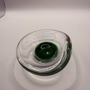 Könnte beinhalten: Eine klare Glasschale mit einem dicken, abgerundeten Rand und einem tiefen, massiven smaragdgrünen kreisförmigen Zentrum. Die Schale hat eine glatte, polierte Oberfläche mit einem schweren Boden, was einen deutlichen Kontrast zwischen den transparenten Außenwänden und dem leuchtend grünen Kern erzeugt.