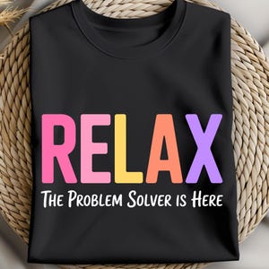 May include: Black t-shirt with the word "RELAX" in pink, yellow, orange, and purple block letters. Below the word "RELAX" is the text "The Problem Solver is Here" in white, all on a black background.