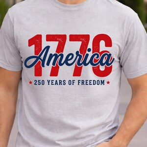 Pu&ograve; includere: T-shirt grigio chiaro con la scritta "1776 America" in rosso e blu. Sotto, c'&egrave; la scritta "250 YEARS OF FREEDOM" con stelle rosse. La maglietta &egrave; girocollo e ha maniche corte.
