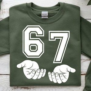 K&ouml;nnte beinhalten: Ein olivgr&uuml;nes Sweatshirt mit der wei&szlig;en Zahl "67". Unter den Zahlen befinden sich zwei wei&szlig;e H&auml;nde mit offenen Handfl&auml;chen. Das Sweatshirt ist von der Marke Gildan.