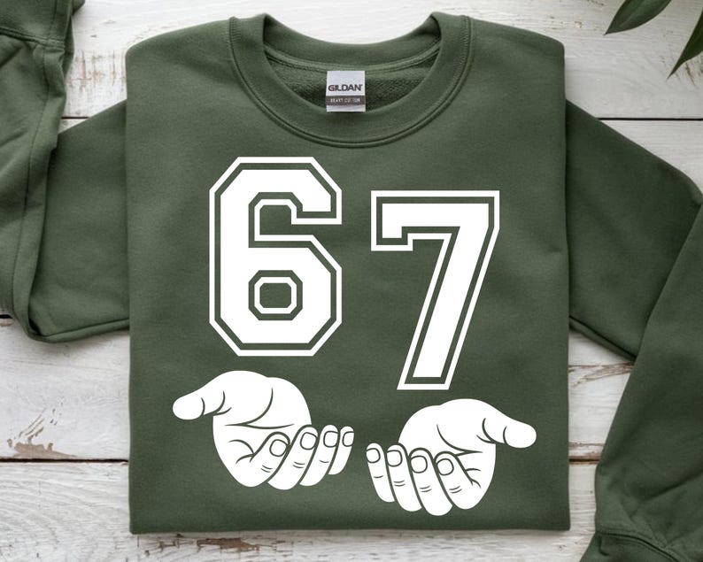 K&ouml;nnte beinhalten: Ein olivgr&uuml;nes Sweatshirt mit der wei&szlig;en Zahl "67". Unter den Zahlen befinden sich zwei wei&szlig;e H&auml;nde mit offenen Handfl&auml;chen. Das Sweatshirt ist von der Marke Gildan.