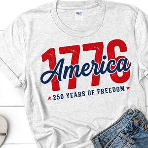 Pu&ograve; includere: Una maglietta grigio chiaro con la scritta "1776 America" in rosso e blu, con "250 YEARS OF FREEDOM" sotto. La maglietta &egrave; piegata e sono visibili un paio di jeans blu e occhiali da sole.