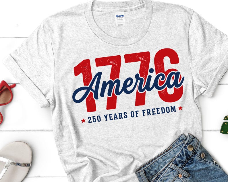 Pu&ograve; includere: Una maglietta grigio chiaro con la scritta "1776 America" in rosso e blu, con "250 YEARS OF FREEDOM" sotto. La maglietta &egrave; piegata e sono visibili un paio di jeans blu e occhiali da sole.
