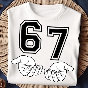 K&ouml;nnte beinhalten: Wei&szlig;es Sweatshirt mit der Zahl 67 in Schwarz mit wei&szlig;em Umriss. Unter den Zahlen befinden sich zwei offene H&auml;nde in einer Schwarz-Wei&szlig;-Strichzeichnung. Das Sweatshirt wird auf einer gewebten Oberfl&auml;che pr&auml;sentiert.