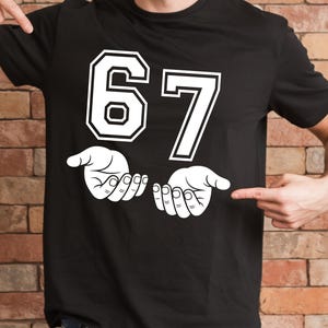 K&ouml;nnte beinhalten: Schwarzes T-Shirt mit der wei&szlig;en Zahl "67", schwarz umrandet. Darunter zwei wei&szlig;e H&auml;nde, Handfl&auml;chen nach oben. Das T-Shirt wird von zwei H&auml;nden gezeigt.