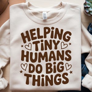 May include: A cream-colored sweatshirt with the phrase "HELPING TINY HUMANS DO BIG THINGS" in brown lettering. The design includes hearts and stars. The sweatshirt is folded on a surface with a succulent and jeans.