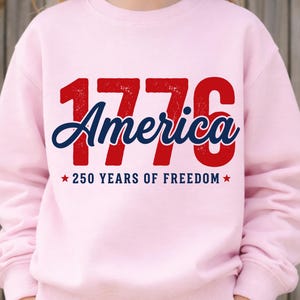 Pu&ograve; includere: Una felpa rosa chiaro con la scritta "1776 America" in rosso e blu, con "250 YEARS OF FREEDOM" sotto. La felpa &egrave; di colore unito e ha un girocollo.