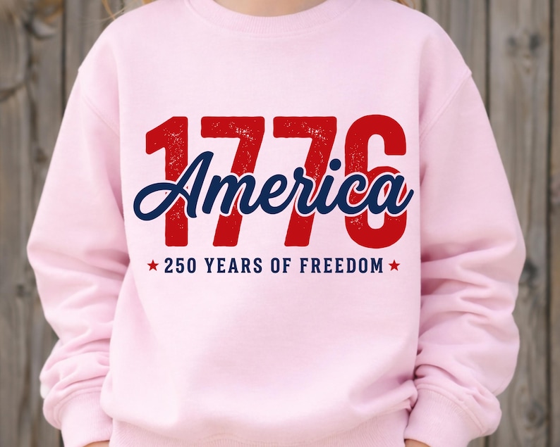 Pu&ograve; includere: Una felpa rosa chiaro con la scritta "1776 America" in rosso e blu, con "250 YEARS OF FREEDOM" sotto. La felpa &egrave; di colore unito e ha un girocollo.