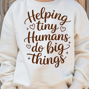 Op de afbeelding: Crèmekleurige sweatshirt met de bruine tekst "Helping tiny Humans do big things". De tekst bevat hart- en steraccenten. De sweatshirt heeft lange mouwen en een ronde hals.