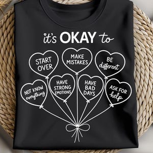 Könnte beinhalten: Schwarzes T-Shirt mit weißem Text und herzförmigen Ballons. Der Text lautet "it's OKAY to" über Ballons mit Sätzen wie "Start Over", "Make Mistakes" und "Be Different". Das Design beinhaltet eine Schleife.