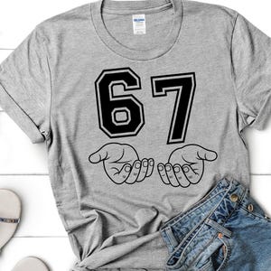 Pode incluir: Camiseta cinza com o n&uacute;mero "67" em preto, contornado em branco, acima de duas m&atilde;os abertas. As mangas est&atilde;o dobradas. &Oacute;culos de sol vermelhos, sand&aacute;lias e jeans vis&iacute;veis.