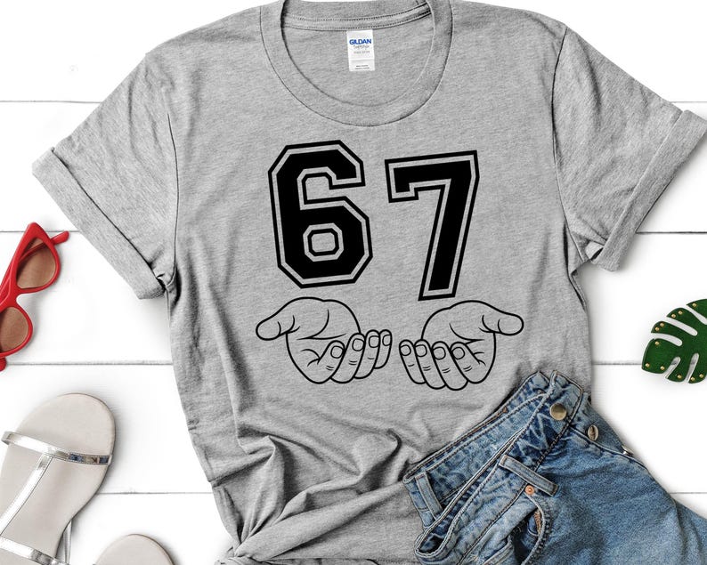 Pode incluir: Camiseta cinza com o n&uacute;mero "67" em preto, contornado em branco, acima de duas m&atilde;os abertas. As mangas est&atilde;o dobradas. &Oacute;culos de sol vermelhos, sand&aacute;lias e jeans vis&iacute;veis.