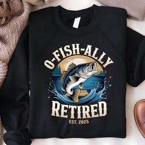 Puede incluir: Sudadera negra con un gr&aacute;fico de un pez saltando fuera del agua. El texto "O-Fish-Ally Retired Est. 2025" est&aacute; impreso en la sudadera.