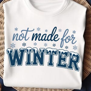May include: White sweatshirt with the phrase "not made for WINTER" in blue script and block letters. Snowflakes are scattered around the text. The sweatshirt is displayed on a woven basket.