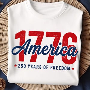 Pu&ograve; includere: T-shirt bianca girocollo con la scritta "1776 America" in rosso e blu. Il testo "250 YEARS OF FREEDOM" &egrave; in basso, con stelle rosse su entrambi i lati. La maglietta &egrave; esposta su una superficie tessuta.