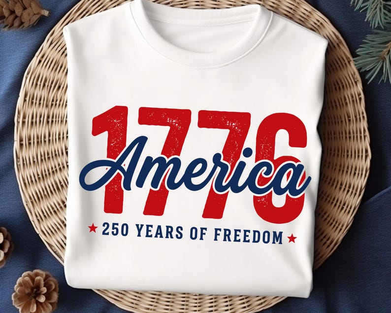 Pu&ograve; includere: T-shirt bianca girocollo con la scritta "1776 America" in rosso e blu. Il testo "250 YEARS OF FREEDOM" &egrave; in basso, con stelle rosse su entrambi i lati. La maglietta &egrave; esposta su una superficie tessuta.