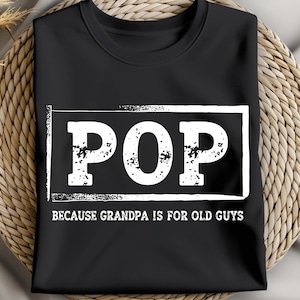 May include: Black t-shirt with the word "POP" in a white, distressed font inside a white rectangle. Below the rectangle, the text reads "BECAUSE GRANDPA IS FOR OLD GUYS". The shirt is displayed on a woven, light brown surface.