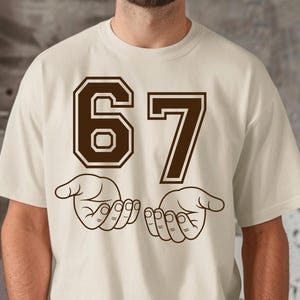 Pode incluir: Camiseta creme com o n&uacute;mero "67" em marrom, com contorno. Abaixo, duas m&atilde;os abertas em estilo de desenho linear. A camiseta &eacute; uma pe&ccedil;a casual e unissex.