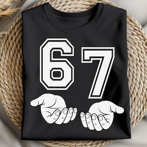 K&ouml;nnte beinhalten: Schwarzes T-Shirt mit der wei&szlig;en Zahl 67 in einer kr&auml;ftigen Schriftart im College-Stil. Darunter befinden sich zwei wei&szlig;e H&auml;nde mit nach oben gerichteten Handfl&auml;chen. Das T-Shirt wird auf einer gewebten, kreisf&ouml;rmigen Oberfl&auml;che pr&auml;sentiert.