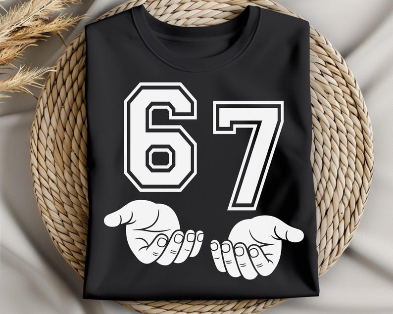 K&ouml;nnte beinhalten: Schwarzes T-Shirt mit der wei&szlig;en Zahl 67 in einer kr&auml;ftigen Schriftart im College-Stil. Darunter befinden sich zwei wei&szlig;e H&auml;nde mit nach oben gerichteten Handfl&auml;chen. Das T-Shirt wird auf einer gewebten, kreisf&ouml;rmigen Oberfl&auml;che pr&auml;sentiert.