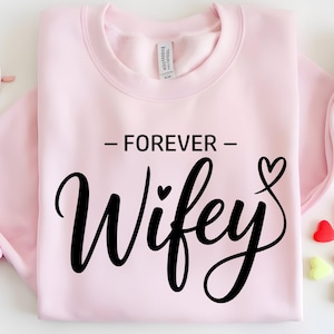 May include: A light pink sweatshirt with the words "- FOREVER - Wifey" in black script. A heart graphic is next to the word "Wifey". The sweatshirt is folded, with a tag visible at the neckline. Heart-shaped candies and sunglasses are in the background.
