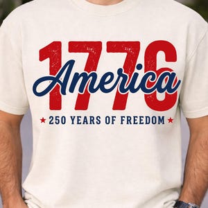 Pu&ograve; includere: T-shirt color crema con la scritta "1776 America" in rosso e blu. Sotto, "250 YEARS OF FREEDOM" con stelle rosse. La maglietta &egrave; in stile casual.