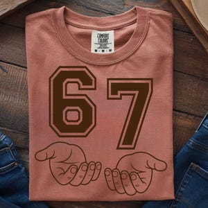 Pode incluir: Camiseta rosa empoeirado com o n&uacute;mero "67" em marrom escuro, com uma fonte em estilo universit&aacute;rio. Abaixo dos n&uacute;meros, duas m&atilde;os abertas em um desenho de linha. A camiseta est&aacute; dobrada e exposta em uma superf&iacute;cie de madeira.