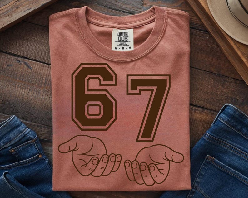 Pode incluir: Camiseta rosa empoeirado com o n&uacute;mero "67" em marrom escuro, com uma fonte em estilo universit&aacute;rio. Abaixo dos n&uacute;meros, duas m&atilde;os abertas em um desenho de linha. A camiseta est&aacute; dobrada e exposta em uma superf&iacute;cie de madeira.