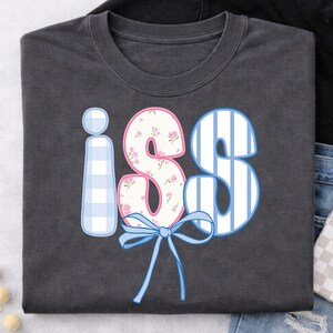 以下が含まれることがあります： 遊び心のあるデザインの「iS」という文字が入った濃いグレーのTシャツ。「i」は水色のギンガムチェック、「S」は白と青のストライプ、「S」はピンクのアクセントの花柄。下には青いリボンがあります。