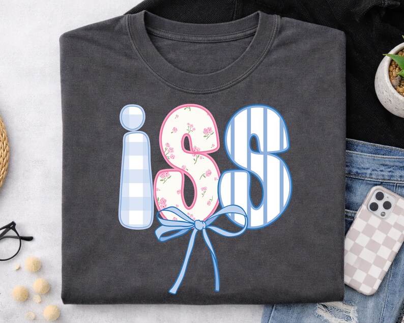 以下が含まれることがあります： 遊び心のあるデザインの「iS」という文字が入った濃いグレーのTシャツ。「i」は水色のギンガムチェック、「S」は白と青のストライプ、「S」はピンクのアクセントの花柄。下には青いリボンがあります。