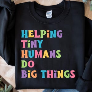 May include: Black sweatshirt with the text "HELPING TINY HUMANS DO BIG THINGS" in rainbow colours.