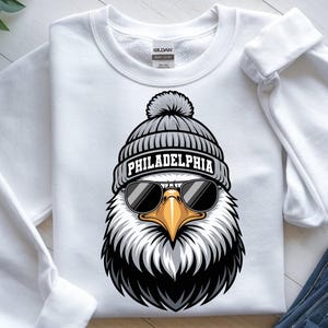 Könnte beinhalten: Weißes Sweatshirt mit einer Grafik eines Adlers, der eine graue Mütze mit dem Wort "PHILADELPHIA" und Sonnenbrille trägt. Der Adler hat einen gelben Schnabel und schwarz-weiße Federn. Das Sweatshirt ist aus weichem Material.