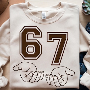 Pode incluir: Camisola creme com o n&uacute;mero 67 em castanho, com uma fonte de estilo universit&aacute;rio. Abaixo dos n&uacute;meros, duas m&atilde;os abertas em contorno castanho. Uma pe&ccedil;a de roupa casual e confort&aacute;vel.